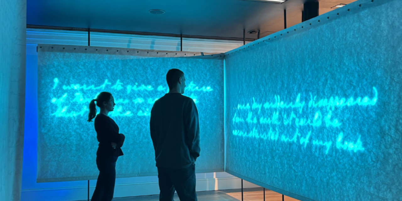 Big blue board with illuminated writing in script font with two people stood in front admiring it