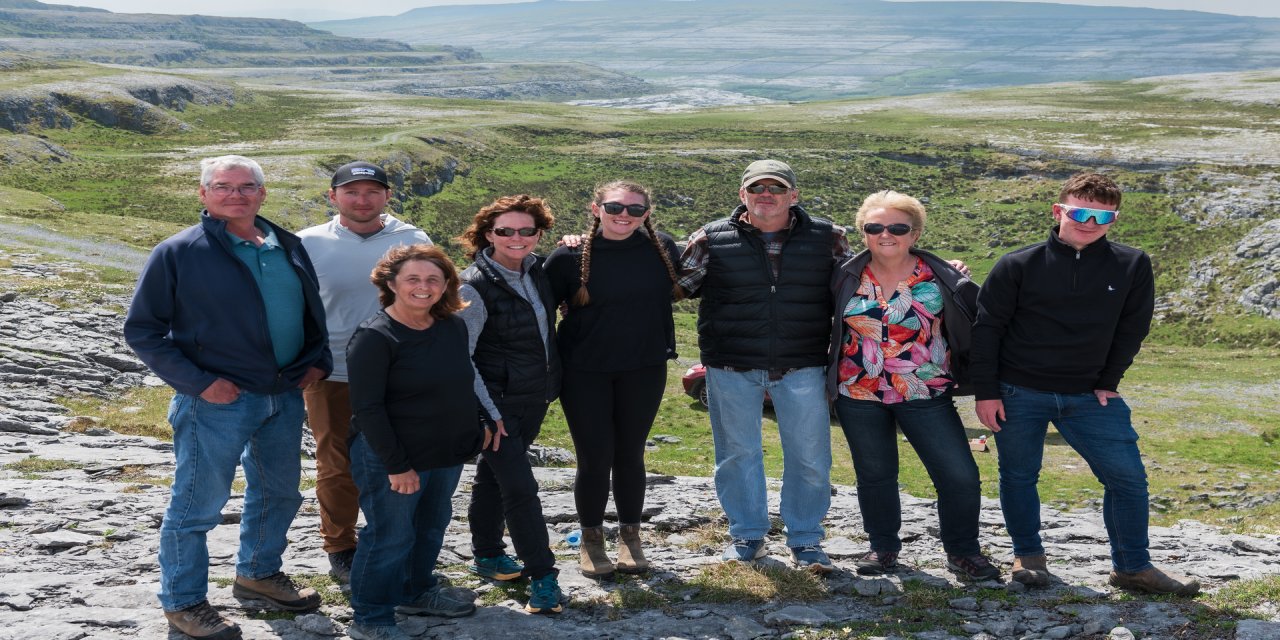 Group photo of a tourist group standing on the side of the Burren Mountains, smiling and posing together. The ground is full of grey cobble stone while the background views is full of greenery. 