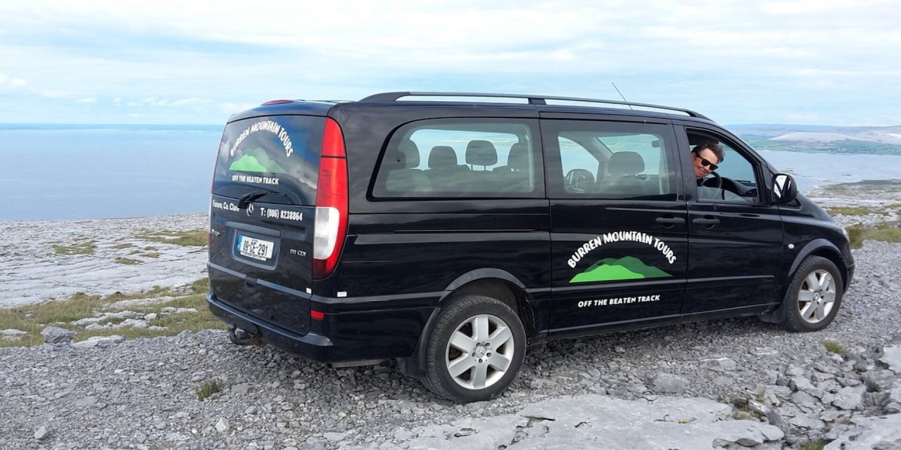 Black, branded van with Burren Mountain stuck onto the side door. Black van parked on the edge of a cliff with view of blue water in the background. Man wearing glasses, smiling with his head poked out of the driver's window. 