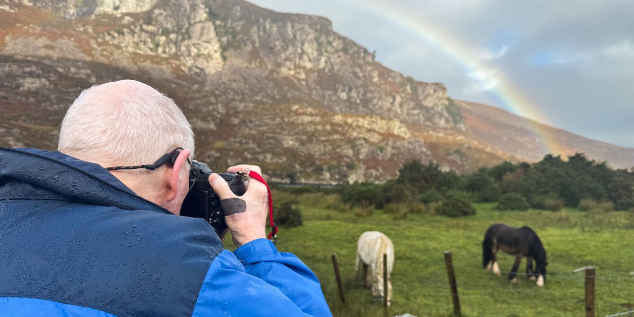 Picture of a man taking a photo of two horses in a field with a mountain and a rainbow in the background