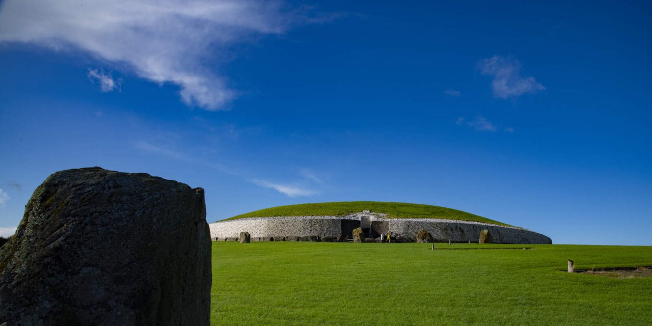 Bright blue skies with small white fluffy cloud in the top left corner. Bright Green grass with stone and grass historic site on the hill.