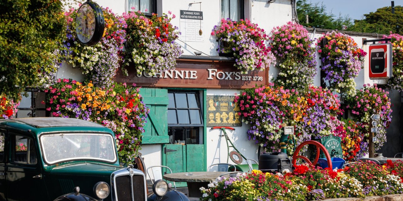 White building covered in multi-coloured flowers. Bright green wooden door with green old car parked outside of the Johnnie Fox's Pub